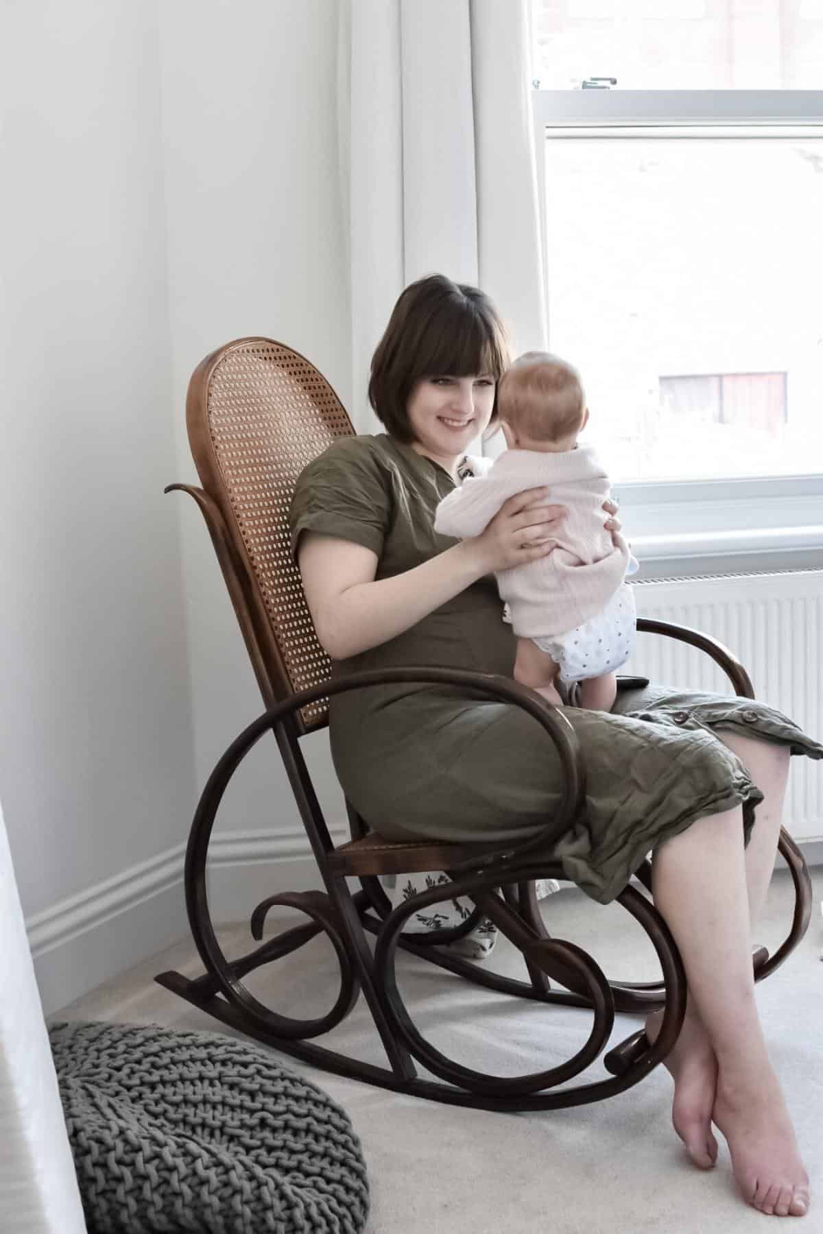 A woman, gently rocking in a wooden chair, holds her baby in a softly lit room. Smiling down at the baby dressed in light attire, she nestles near a knitted gray cushion. Natural light streams through a large window, adding warmth to this serene moment.
