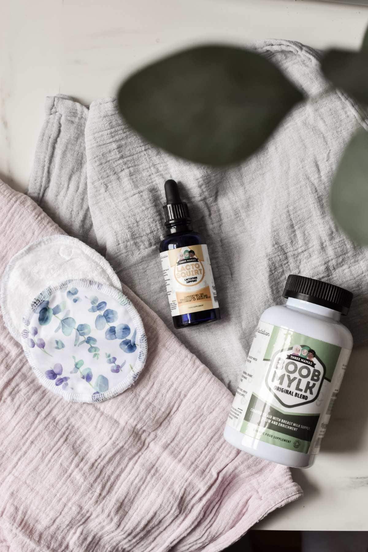 A flat lay of a cloth fabric surface showcases the floral-patterned reusable pad alongside a blue dropper bottle labeled Postpartum Balance and a white jar labeled Boob Mylk Lactation Blend from Three Mamas Natural, surrounded by soft fabrics. Perfect for new mothers seeking breastfeeding supplements.
