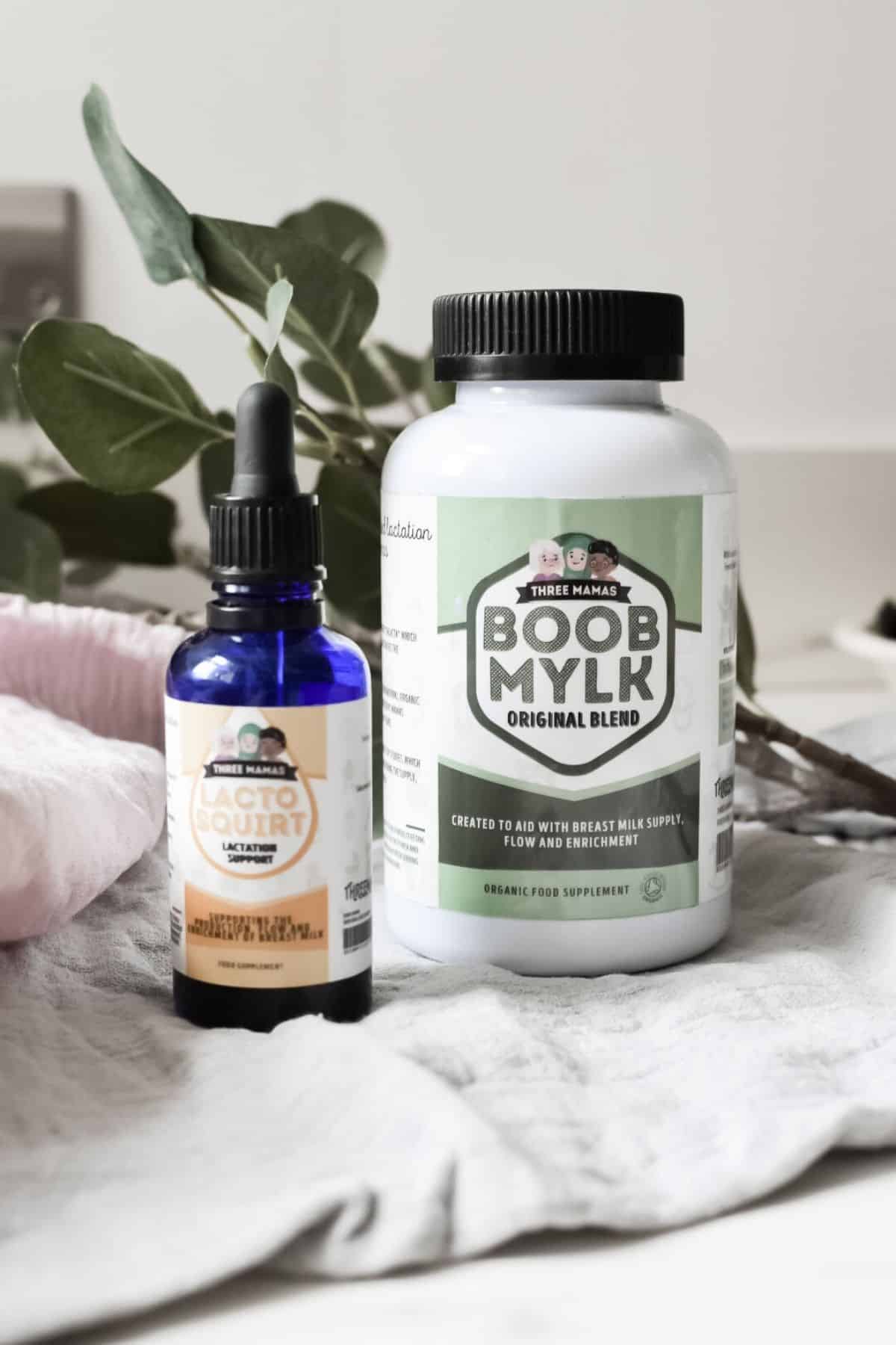 A bottle of Natura Lactation Goat Squirt sits beside a container of Three Hams Boob Mylk Original Blend, with fresh green leaves in the backdrop. These Natural Breastfeeding Supplements are crafted to aid lactation and breastfeeding by Three Mamas.