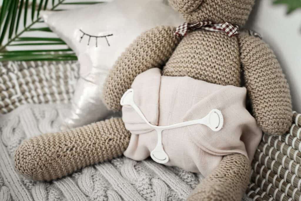 A knitted teddy bear wearing a soft pink budget muslin cloth nappy sits on a textured gray blanket. Behind it, a plush star pillow with embroidered closed eyes adds charm, while a green plant frond peeks out. 