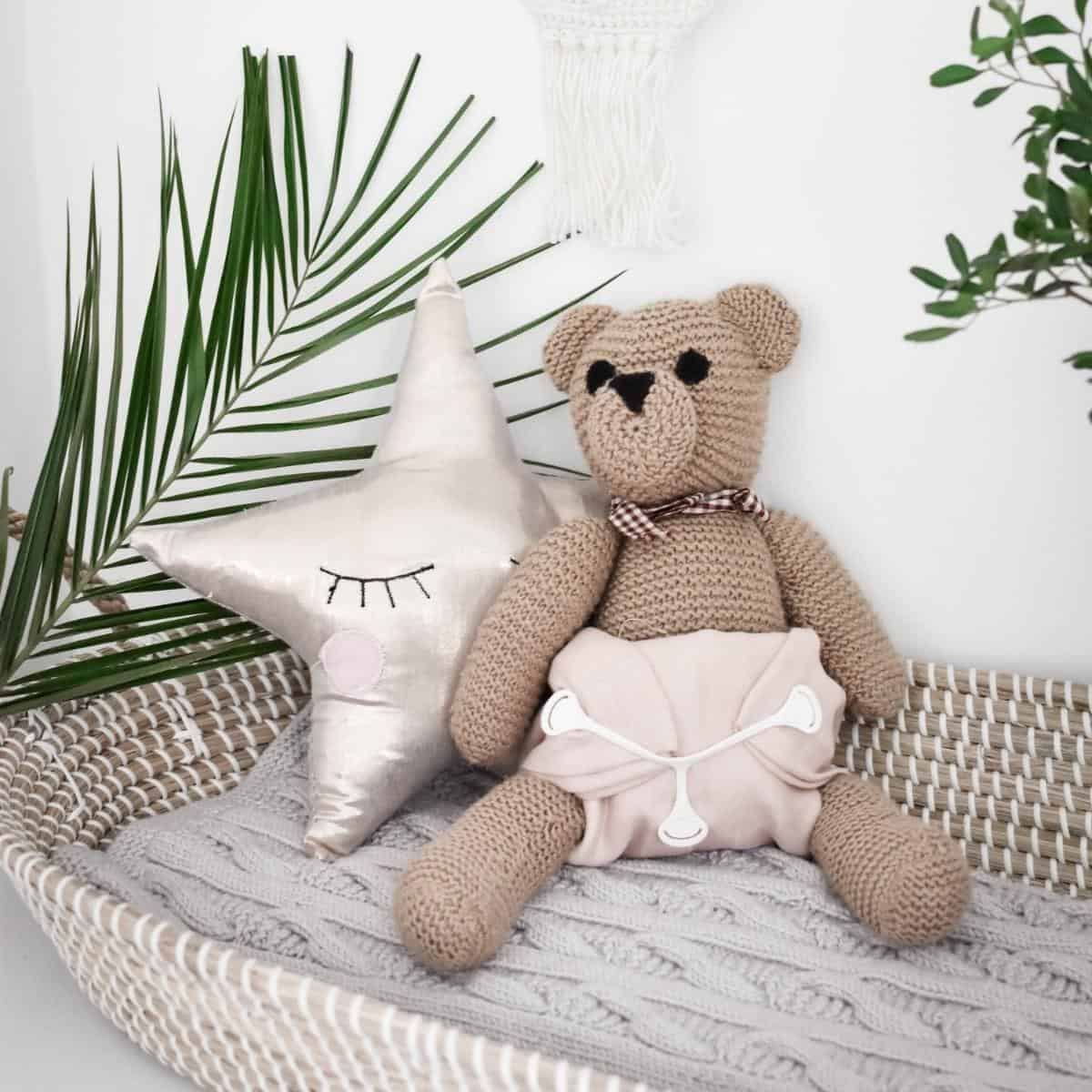 A knitted teddy bear wearing a muslin cloth nappy sits in a woven basket next to a smiling star-shaped pillow. Green palm fronds are in the background, adding a touch of nature to the cozy setup.