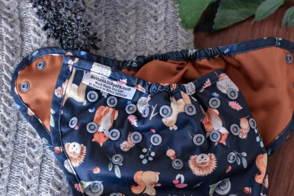 Cloth diaper with a playful animal design, featuring squirrels, hedgehogs, foxes, and lions on a dark background. This adorable Buttons Diapers nappy rests on a knitted gray blanket beside a small cluster of dark berries.