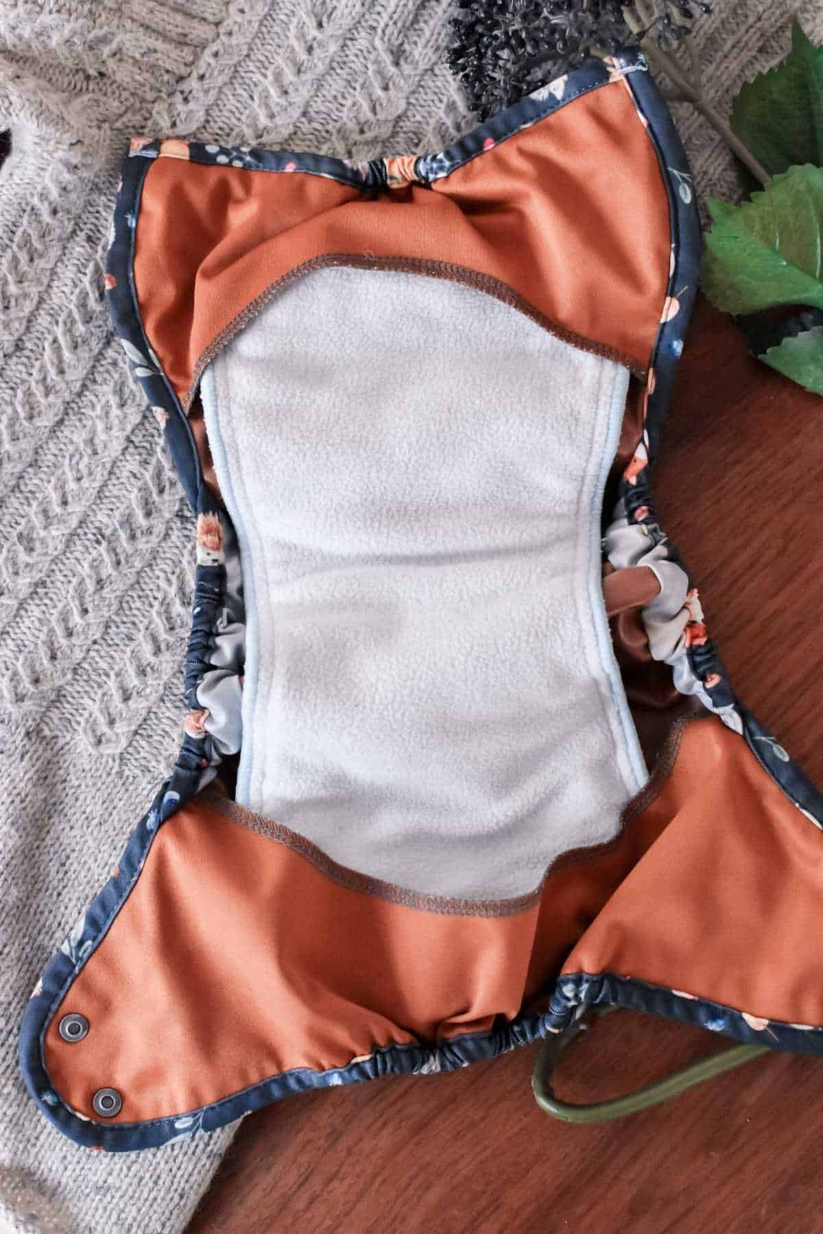 Laid open is a Buttons Diapers reusable cloth diaper, showcasing its white absorbent interior and vibrant orange patterned exterior. It rests on a textured neutral knit surface, while a leafy green plant adds a touch of nature in the background.
