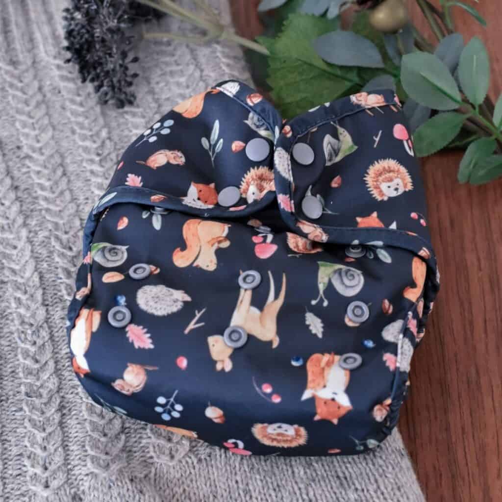A Buttons Diapers reusable cloth diaper showcases woodland animals like foxes, hedgehogs, and squirrels against a dark background. Nestled on knitted gray fabric with green leaves and tiny decorative fruits nearby.