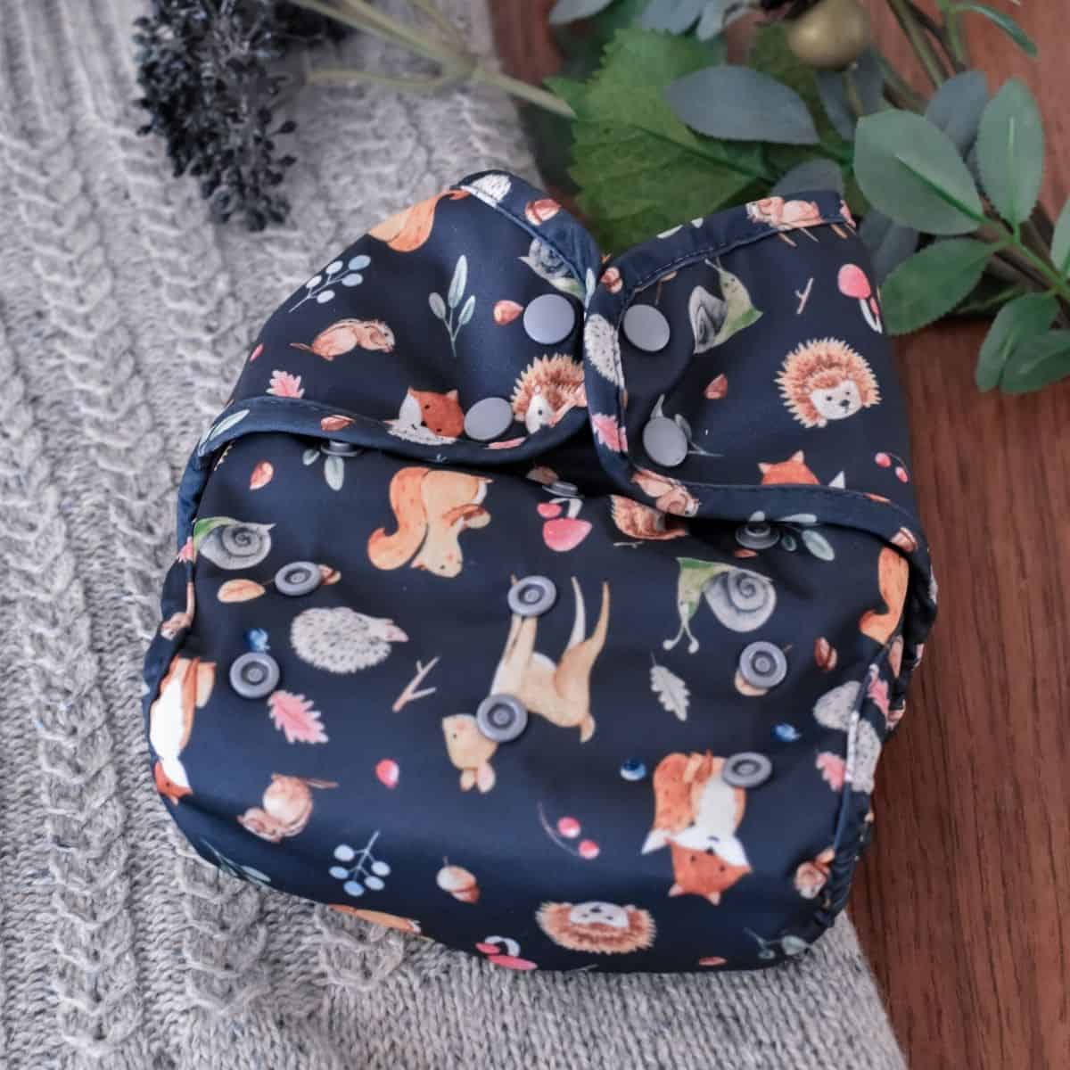 A Buttons Diapers reusable cloth diaper showcases woodland animals like foxes, hedgehogs, and squirrels against a dark background. Nestled on knitted gray fabric with green leaves and tiny decorative fruits nearby.