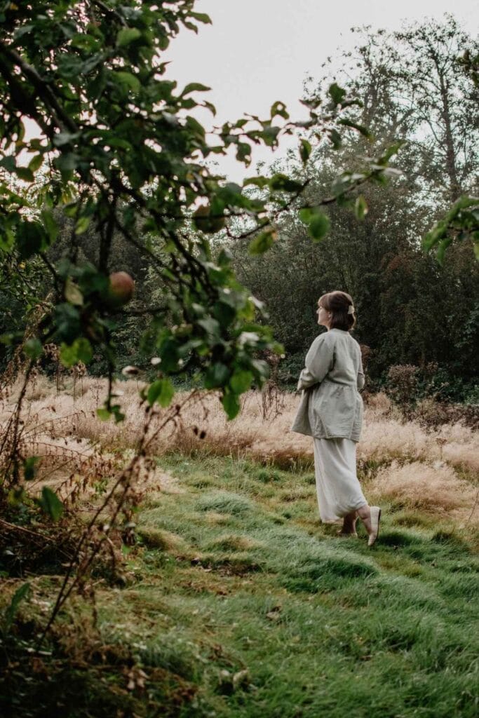 A person in a light outfit stands on a lush green grass path, carefully considering clothing maintenance while gazing at an apple tree with overhanging branches. The background features tall, dried grass and dense trees, creating a peaceful, natural setting.