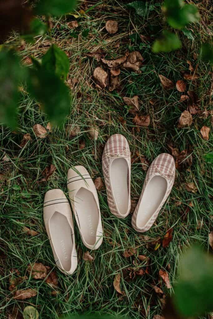 Two pairs of womens slip-on shoes lie on grass surrounded by leaves. On the left, beige shoes with a crisscross design; on the right, plaid-patterned shoes in shades of red and white.