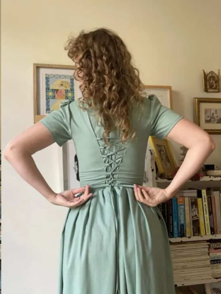 A woman with curly hair wearing a soft green dress with a corset-style lace-up back, adjusting the ties. The dress has a romantic, historical-inspired feel, perfect for cottagecore sewing.