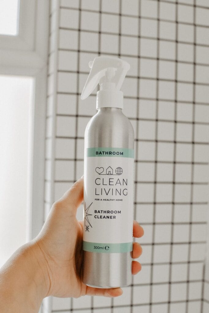 A hand holds a silver spray bottle labeled Clean Living Bathroom Cleaner against a white tiled wall. The label features a heart, house, and leaf icon, underscoring a healthy home concept. With 300ml of natural formula, Clean Living Cleaning Products offer effective and sustainable solutions.