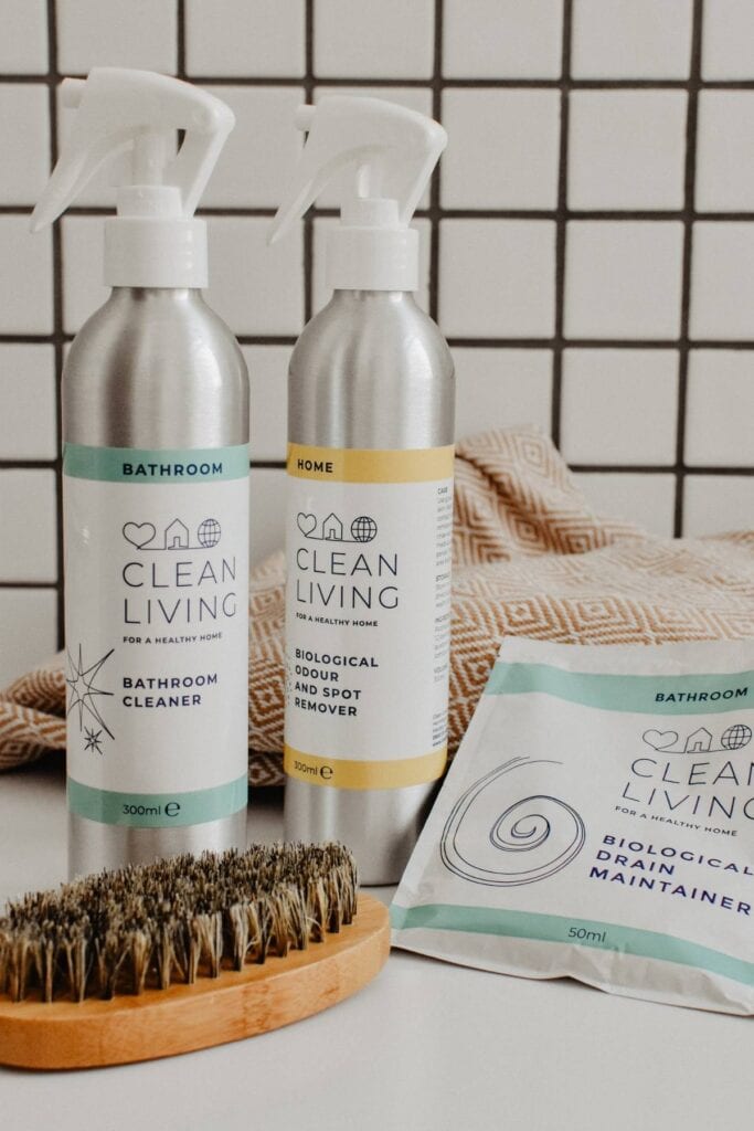 Two Clean Living cleaning product bottles on a surface: a bathroom cleaner with a green label and an odor remover with a yellow label. Nearby are a packet of Natural & Sustainable drain maintainer and a wooden brush. The background features a tiled wall and a beige cloth.