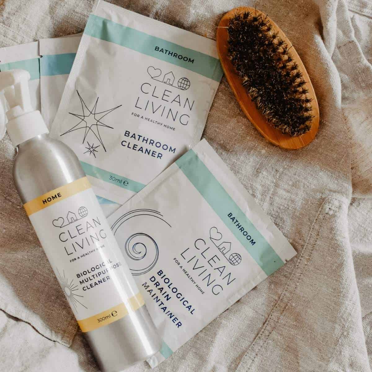 A flat lay image of sustainable, eco-friendly cleaning products on a beige fabric surface. It includes a silver spray bottle, two packets labeled Clean Living Bathroom Cleaner, and a natural bristle brush with a wooden handle.
