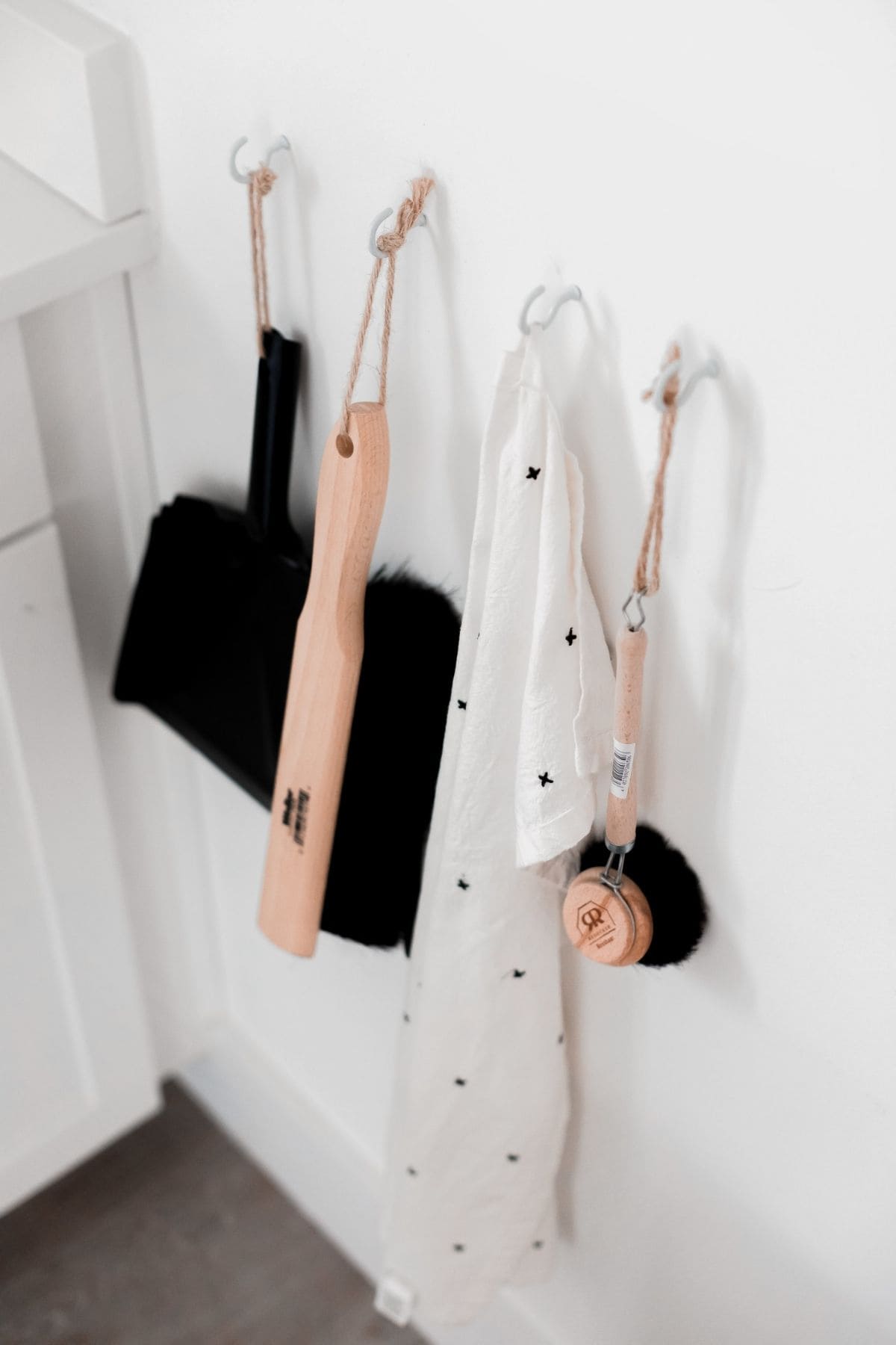 Cleaning tools hang on wall hooks, featuring a black dustpan and brush, a wooden-handled brush, a white cloth with small black stars, and a small wooden strainer. With this organized setup, its easier to stay on top of your cleaning schedule. The wall is white, and the floor is light-colored.