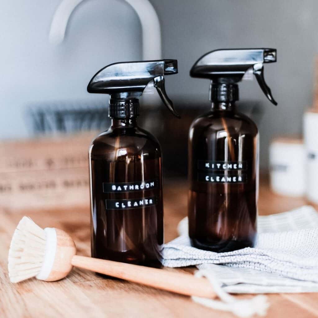 Two brown spray bottles labeled Bathroom Cleaner and Kitchen Cleaner rest on a wooden countertop, accompanied by a scrub brush and striped cloth. A blurred kitchen sink is in the background.