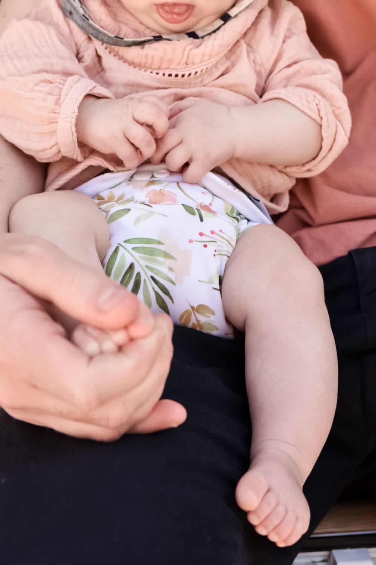 A baby in a floral-patterned cloth nappy sits on a persons lap, clad in a peach outfit. With little fingers touching, the baby enjoys the gentle hold of a hand on their foot.