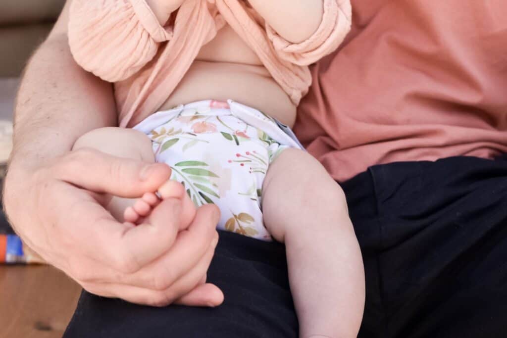 A baby in a floral diaper sits on an adults lap, who could easily be the go-to cloth nappy dad for tips. The scene captures tenderness as the adult, casually dressed in a pink shirt and black pants, gently holds the babys foot.