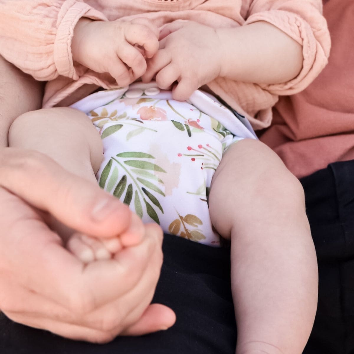 A baby wearing a colorful cloth nappy sits on Dad's lap, who gently holds one of the baby's feet. The close-up scene captures the babys hands and legs against the soft fabric, offering a warm glimpse into lifes little moments.