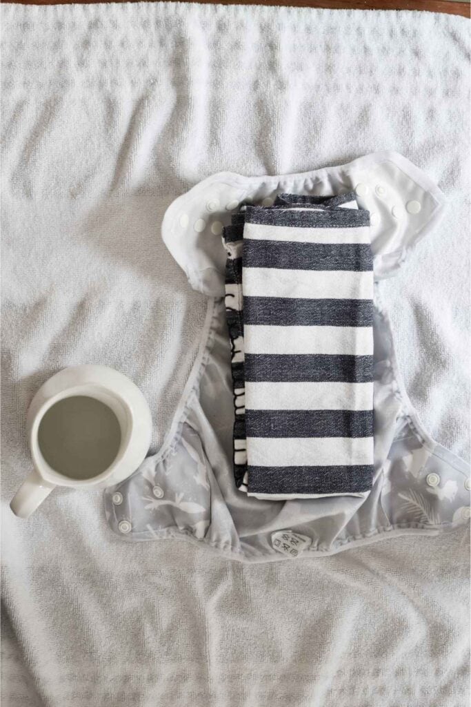 A white cloth nappy lies open on a towel, ready to perform the tea towel test. Inside, a folded black and white striped tea towel is placed. To the left of the diaper sits a white ceramic mug filled with water.