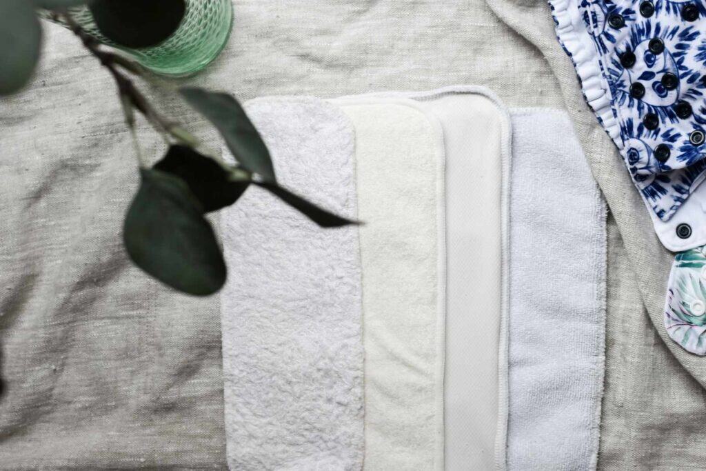 A stack of cloth nappy absorbent inserts in various soft colors rests on a textured fabric background. A leafy branch is partially visible in the upper left corner.