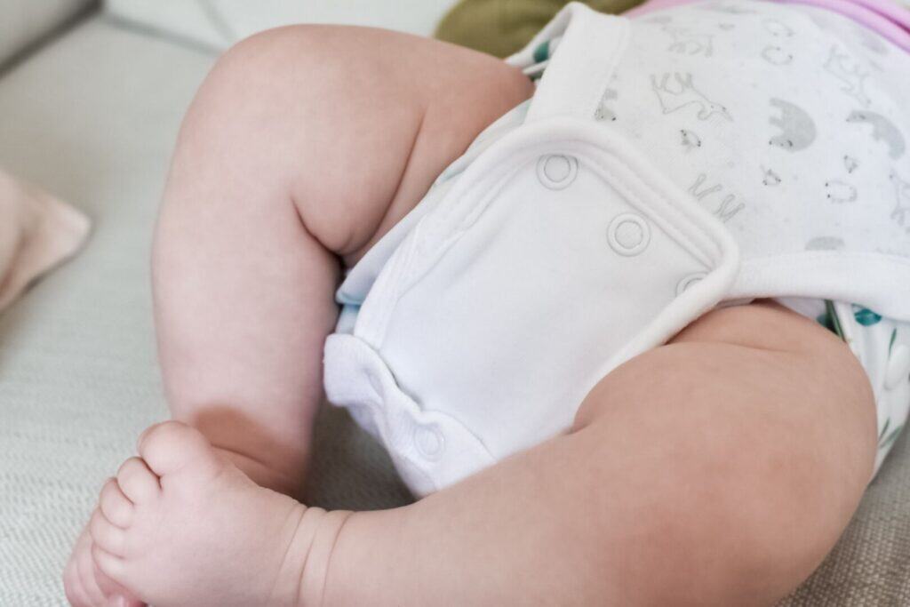A close-up of a babys legs and feet in soft cloth nappies. The baby is wearing a patterned white onesie with a vest extender to help the vest fit over the cloth nappy. 