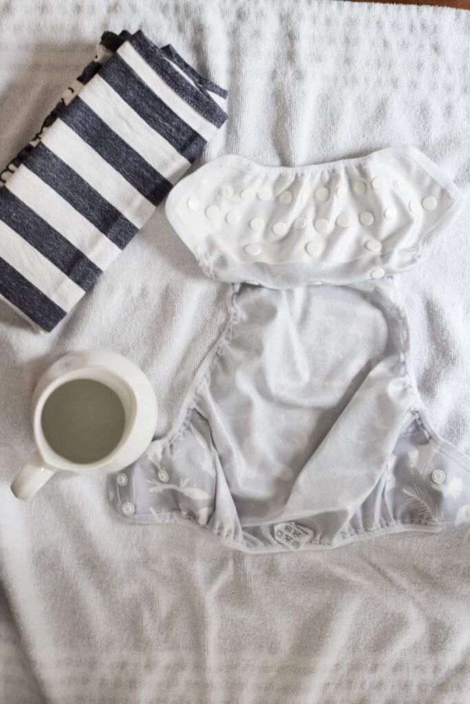 A white cloth nappies is laid open on a towel next to a mug filled with water and a folded blue and white striped cloth.