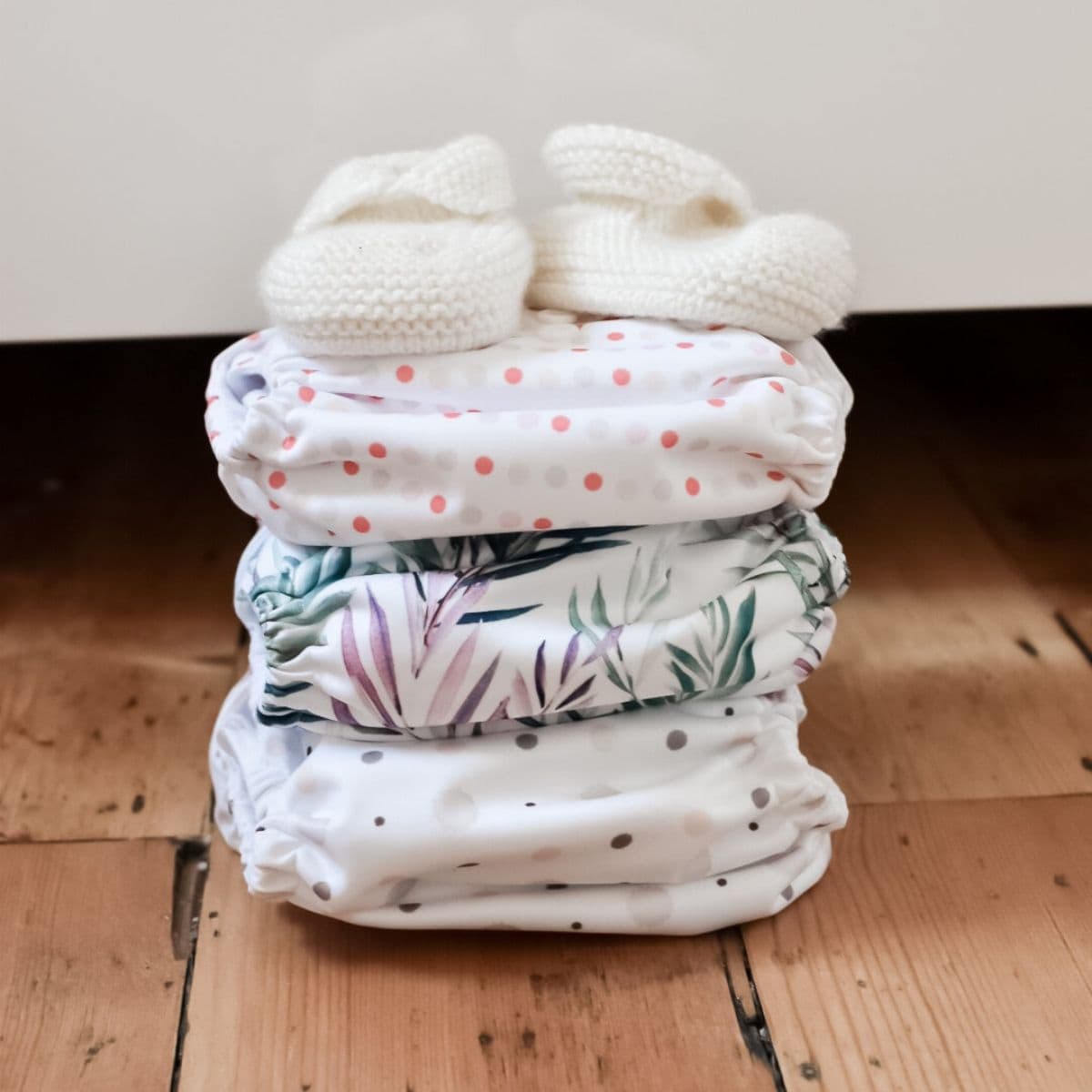 A stack of three patterned cloth nappies is topped with a pair of small white knitted baby booties.