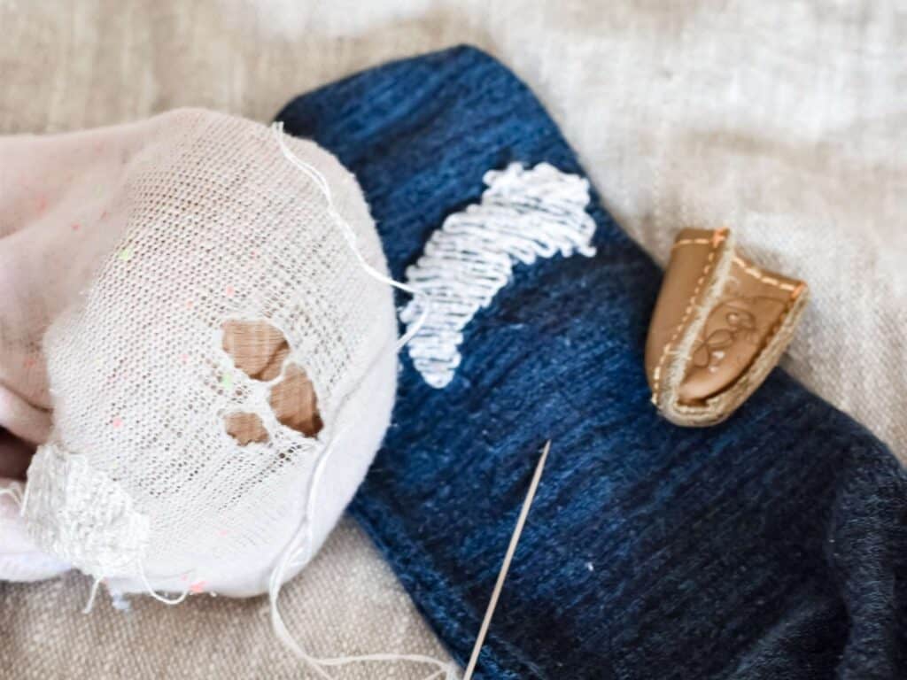 A close-up of a pink and blue sock with visible holes, showing the darning process with a needle and thread in progress.