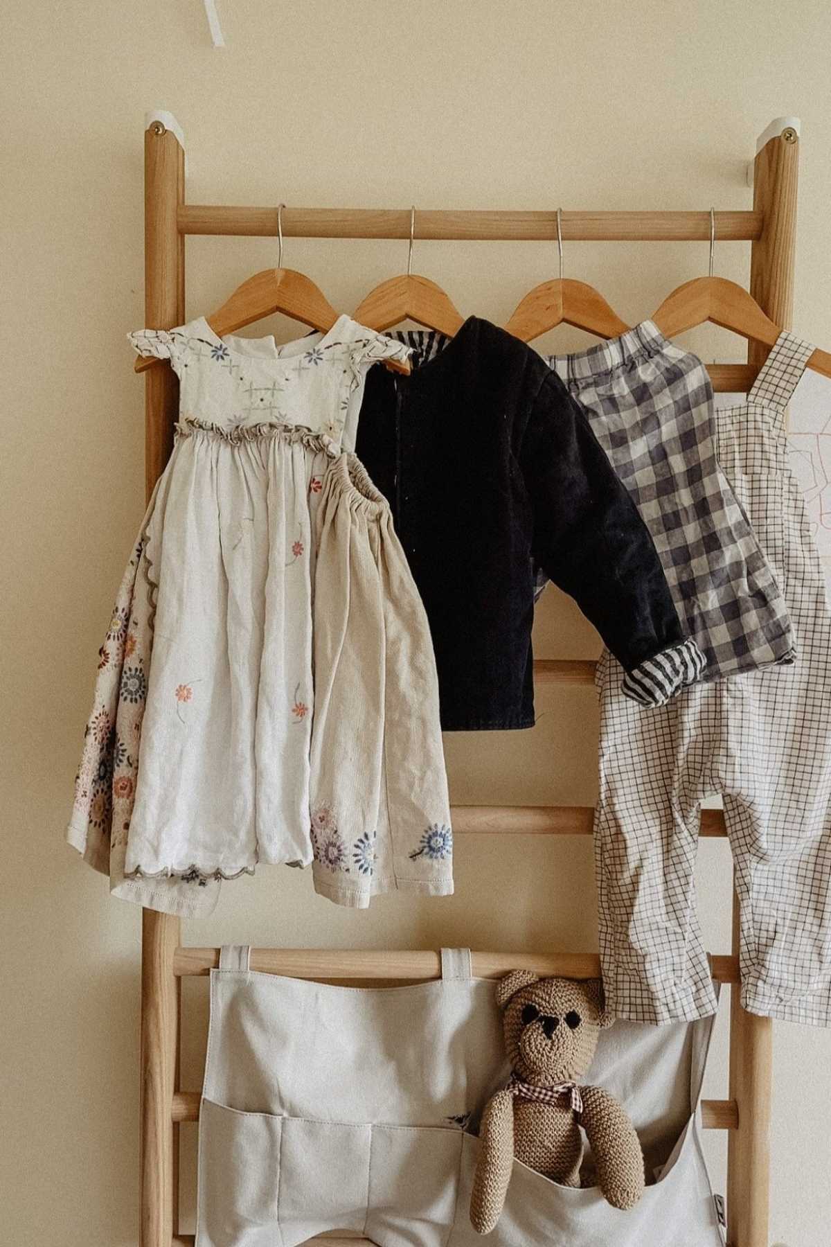 A wooden rack displays three childrens clothing pieces crafted from vintage and deadstock fabrics: a floral dress, a dark jacket, and checkered overalls. Below, a beige fabric organizer holds a small brown stuffed bear in its pocket.