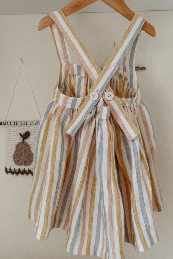 A childs striped dress made from hangs on a wooden hanger. It features vertical pastel stripes in yellow, pink, and blue with cross-back straps fastened by white buttons. In the background, a small decorative wall hanging boasts a crochet pear design.
