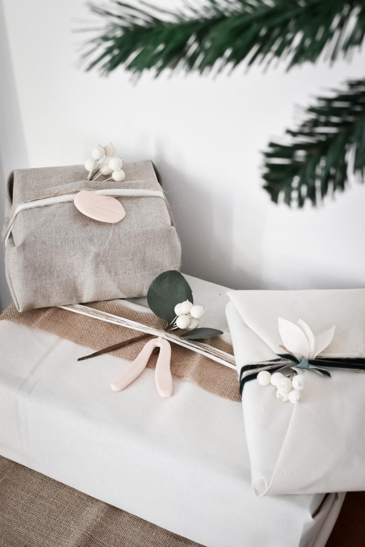 Two elegantly wrapped gifts sit on a table, one in beige fabric and the other in white paper, each adorned with minimalist decorations featuring faux leaves, berries, and DIY clay gift tags. A sprig of a pine tree is partially visible above.