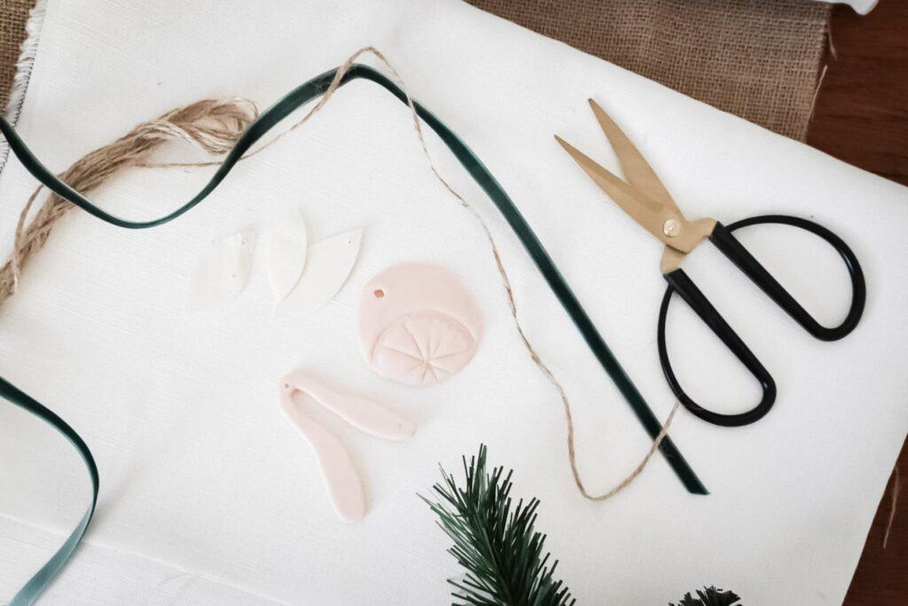 Crafting materials on a white surface: black scissors, green ribbon, and twine perfect for gift wrapping. Small clay shapes resembling citrus slices and leaves are DIY clay gift tags. A piece of green foliage is placed nearby for a natural touch.