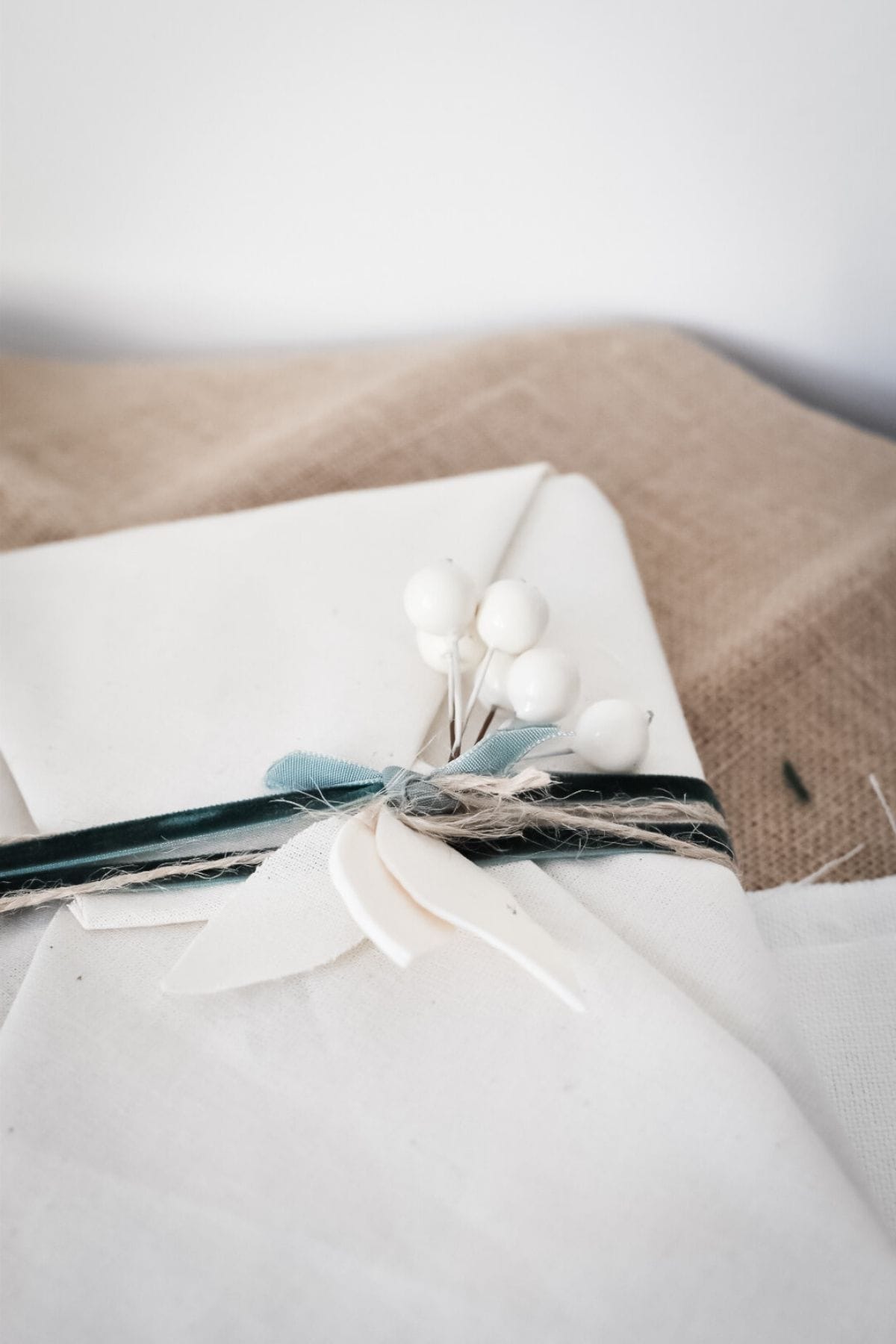 A close-up of a gift, tied with twine and a dark green ribbon, adorned with white berries and a small leaf decoration made from air dry clay.