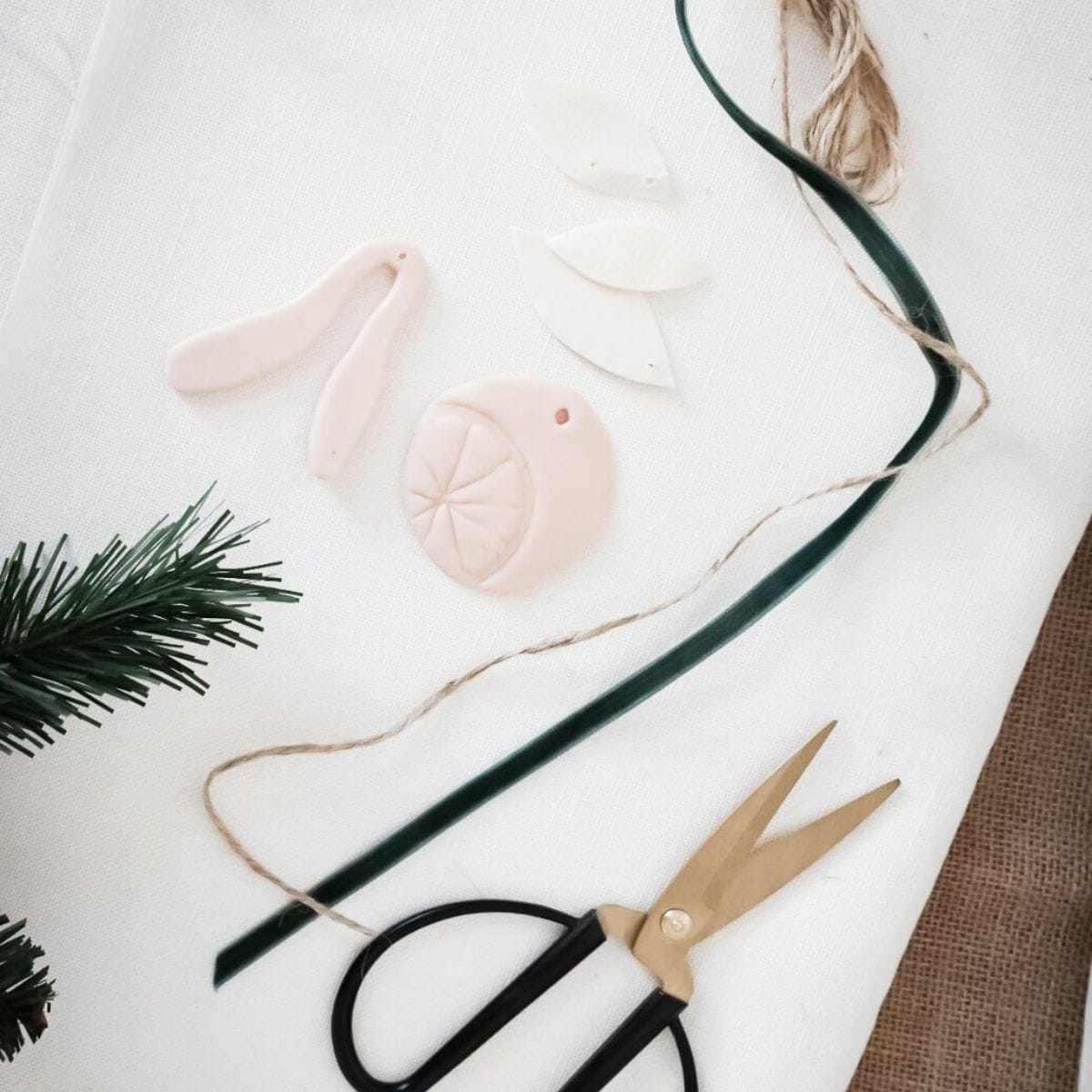 Crafting materials on a white surface include clay shapes like pastel pink tags and citrus-like slices. Scattered nearby are gold scissors, twine, and a strip of green tape.