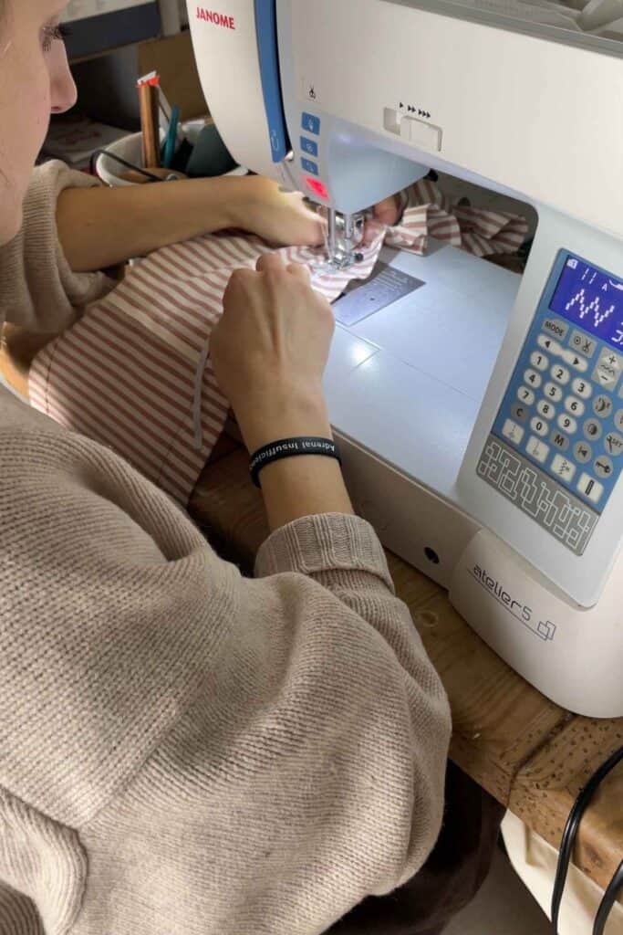 A person uses a sewing machine to stitch a piece of red and white striped fabric. The machine is a Janome atelier 5. 