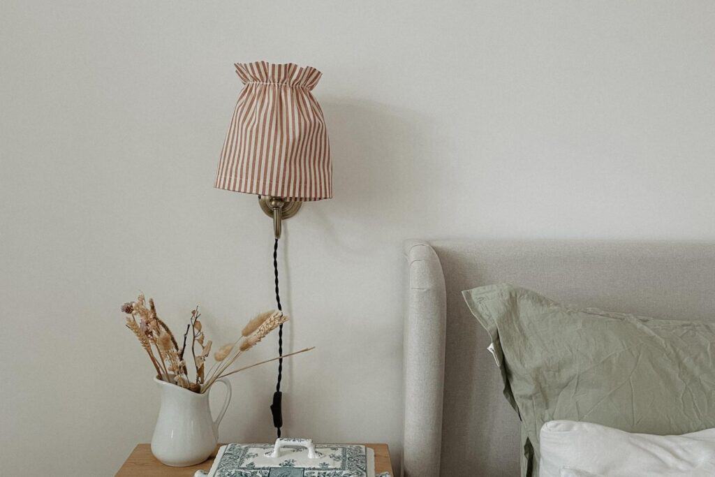 A bedside scene with a striped DIY gathered lampshade on the wall above a nightstand. The nightstand holds a white jug with dried plants, a decorative book, and a tissue box. A pillow with a green pillowcase rests on a light gray bed.