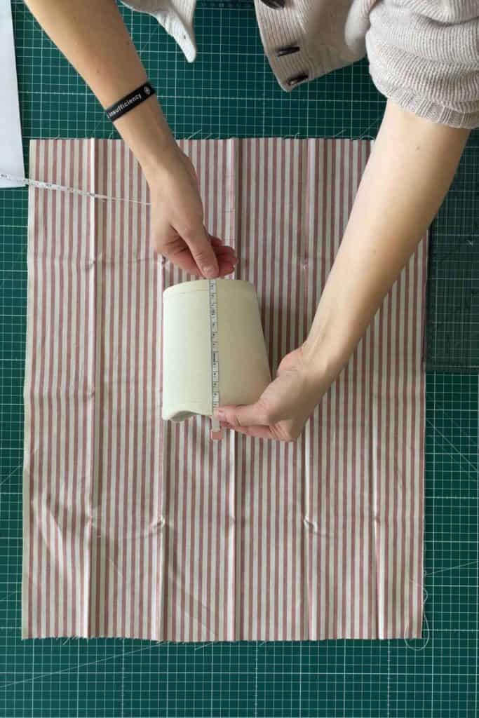 A person measures a lampshade using a measuring tape over a red and white striped fabric on a green grid cutting mat.