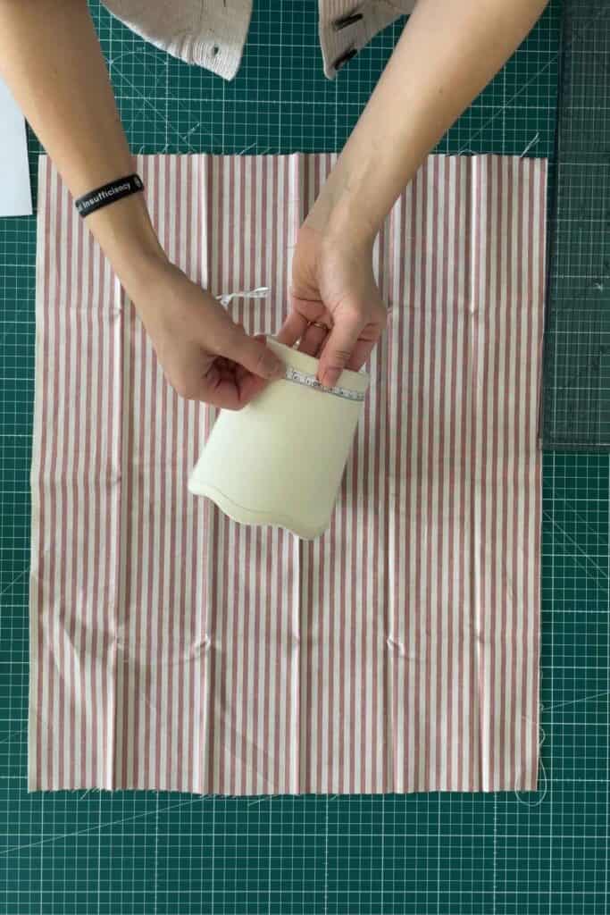 Two hands are measuring a small lampshade, over a larger piece of red and white striped fabric. This is placed on a green cutting mat that has grid lines. A ruler and part of a sweater are visible nearby.