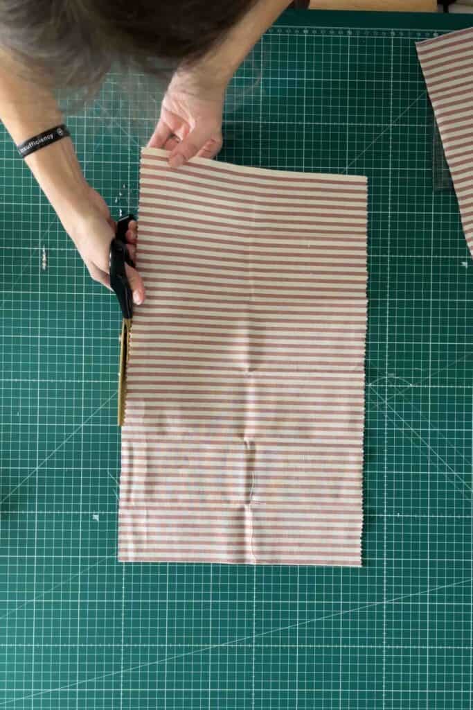 A person is cutting a piece of striped fabric with scissors on a green cutting mat. The fabric has horizontal stripes in red and white. 