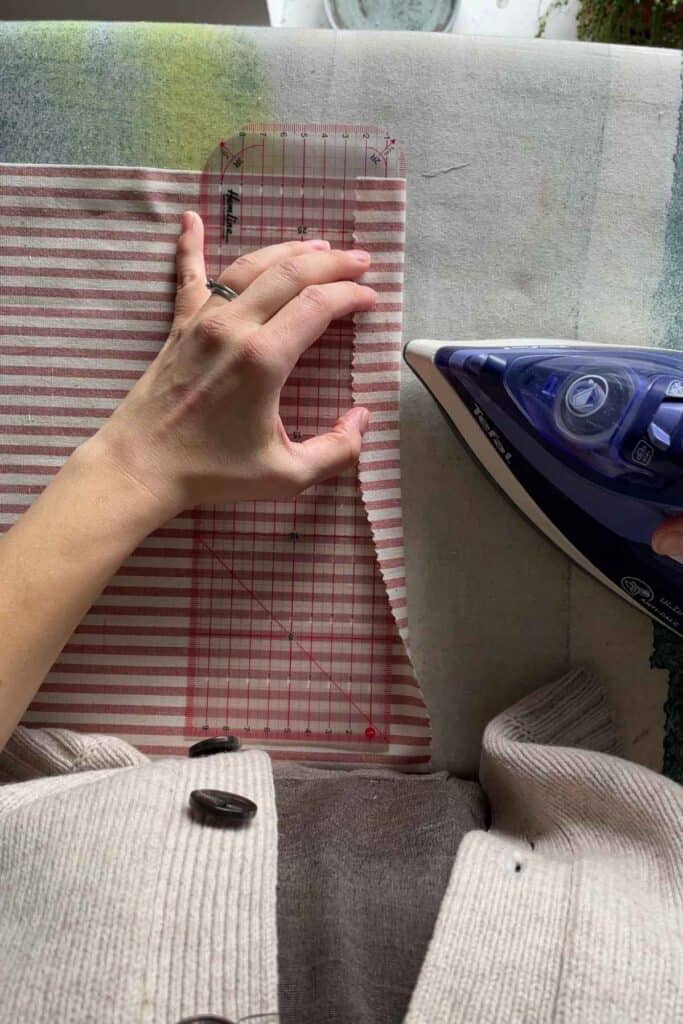 A person wearing a sweater is using an ironing ruler and an iron to press and measure the edge of a piece of red-and-white striped fabric. The fabric is on an ironing board with a slightly worn cover.