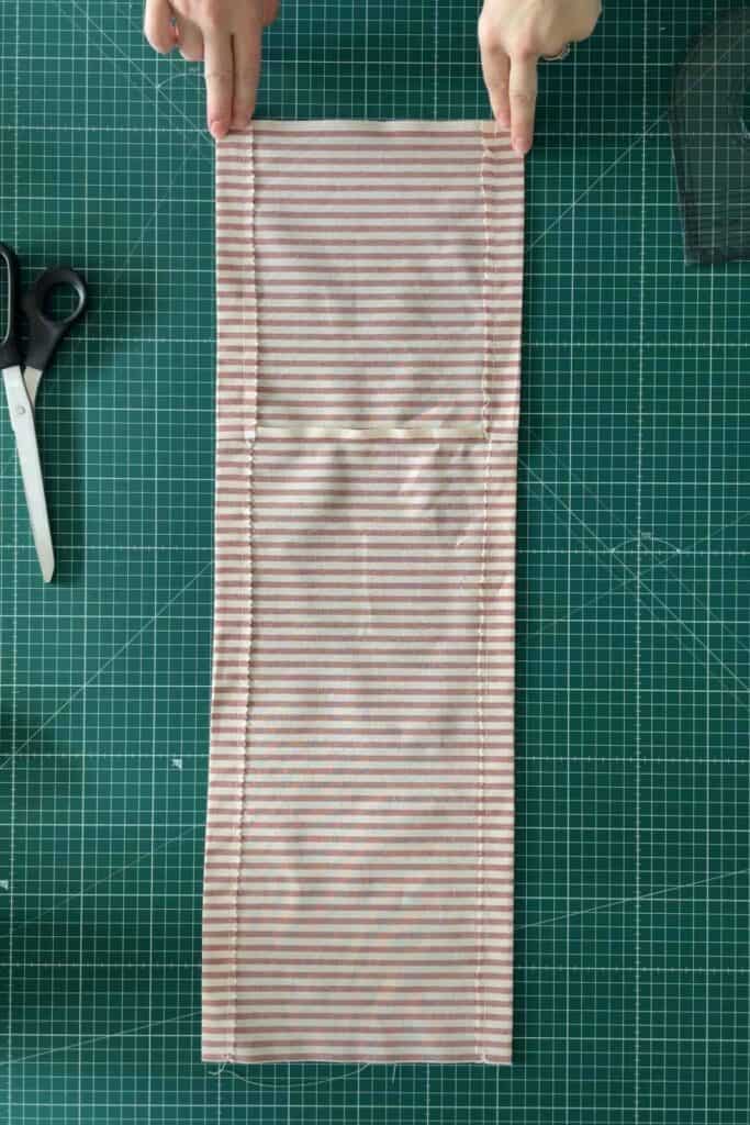 Hands are adjusting a piece of striped fabric on a green cutting mat with a grid pattern. Scissors are placed to the left of the fabric.