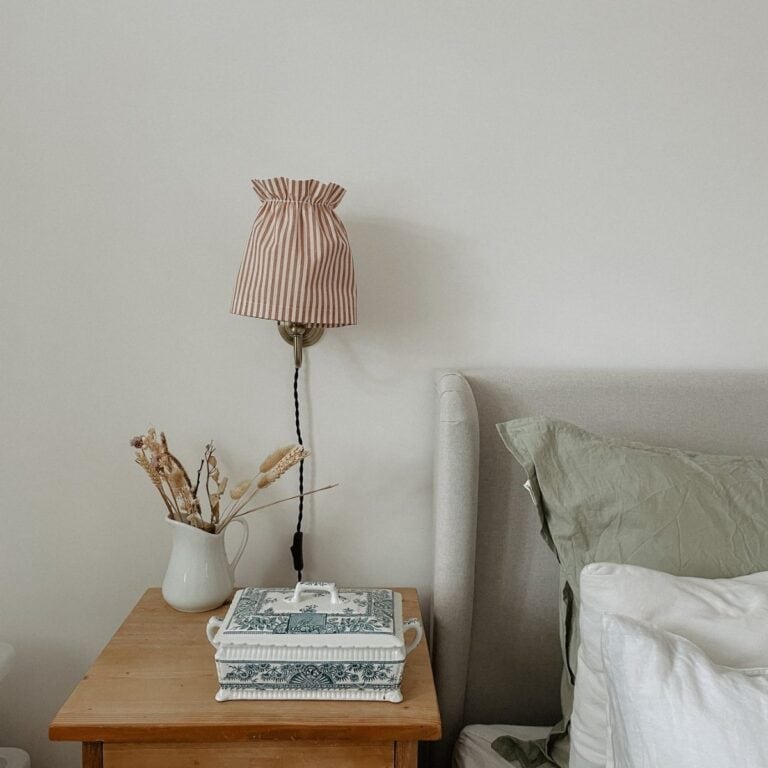 A cozy bedroom nightstand features a striped wall lamp, a green and white lidded floral box, and a white pitcher with dried plants. The neutral-toned bedding has a green pillow and white sheets against a light upholstered headboard.