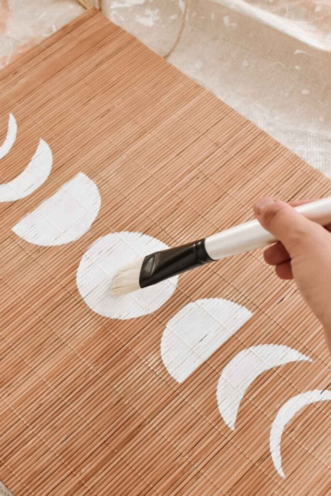 A hand painting white crescent shapes on a woven bamboo mat using a wide brush creates a DIY moon phases motif.