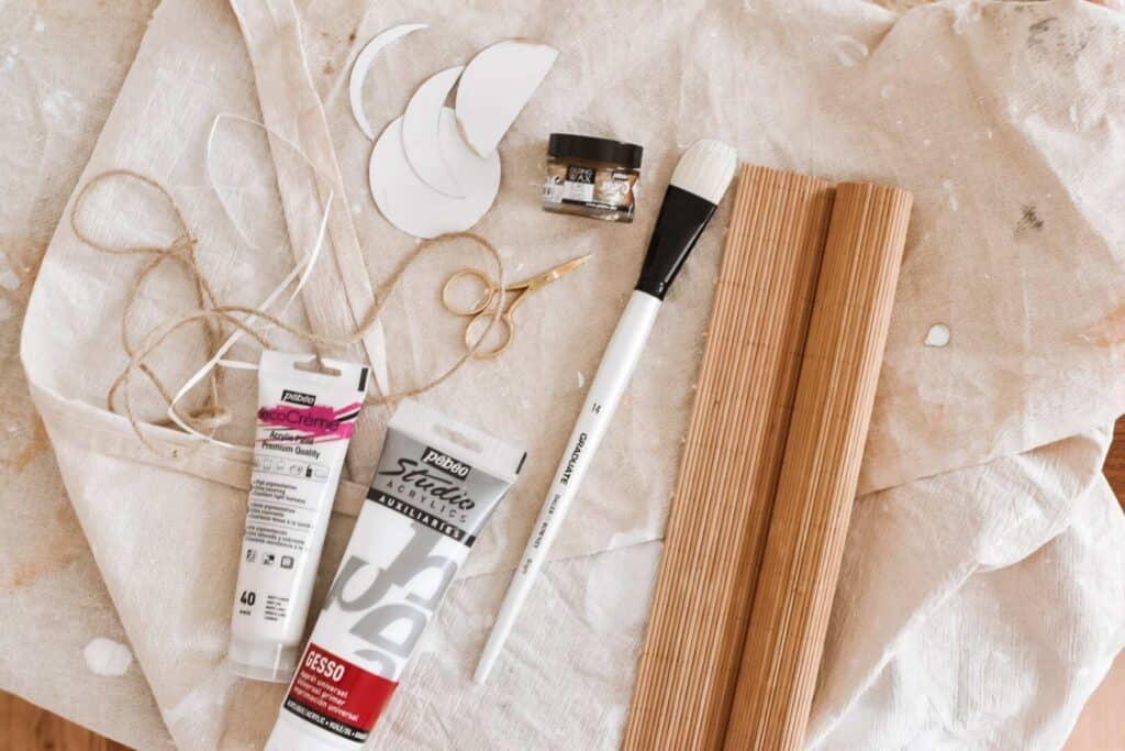 Craft supplies on a cloth surface, featuring tubes of paint, a paintbrush, twine, and materials to create your own DIY moon phases bamboo wall hanging. Included are wooden sticks, a small pair of scissors, white paper cutouts scattered about, and a handy container.