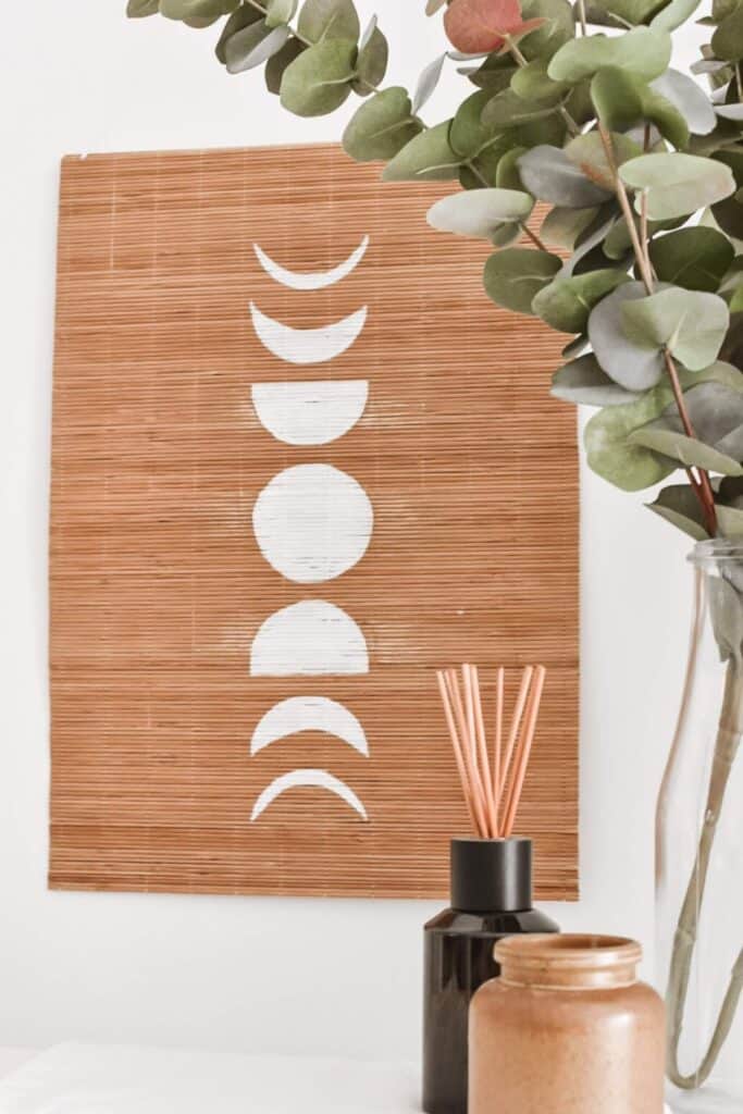 A DIY bamboo wall hanging with white moon phases aligned vertically. Nearby, a vase filled with eucalyptus branches complements the dark reed diffuser and brown jar, all beautifully arranged on a light surface.