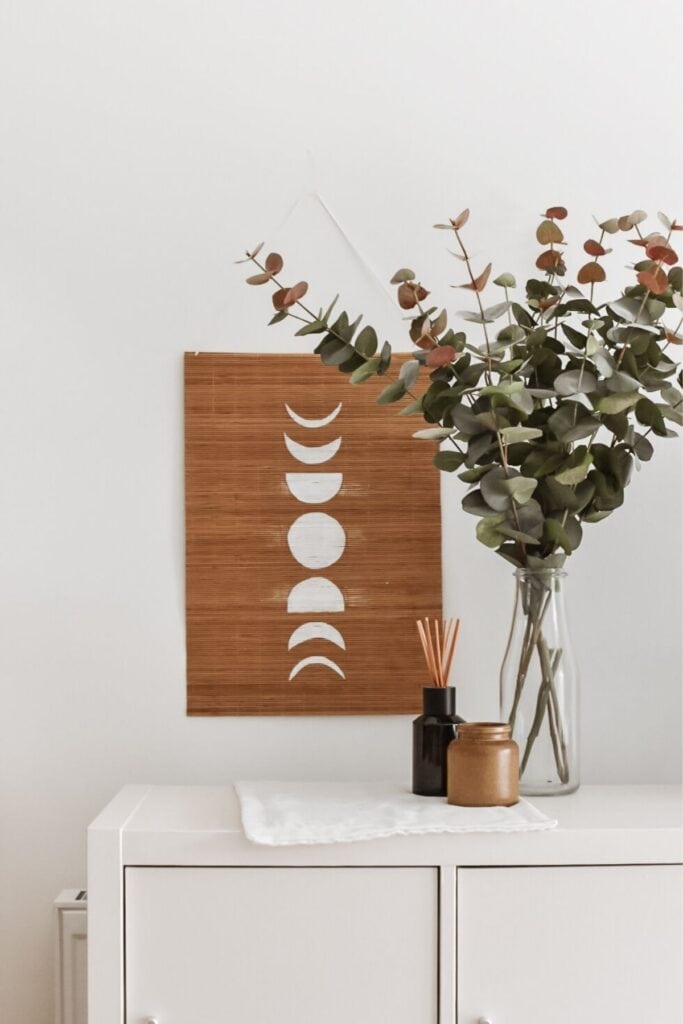 A minimalistic interior features a DIY bamboo moon phase wall art above a white cabinet. A glass vase with eucalyptus and a brown jar with reed diffusers enhance the serene, natural aesthetic.