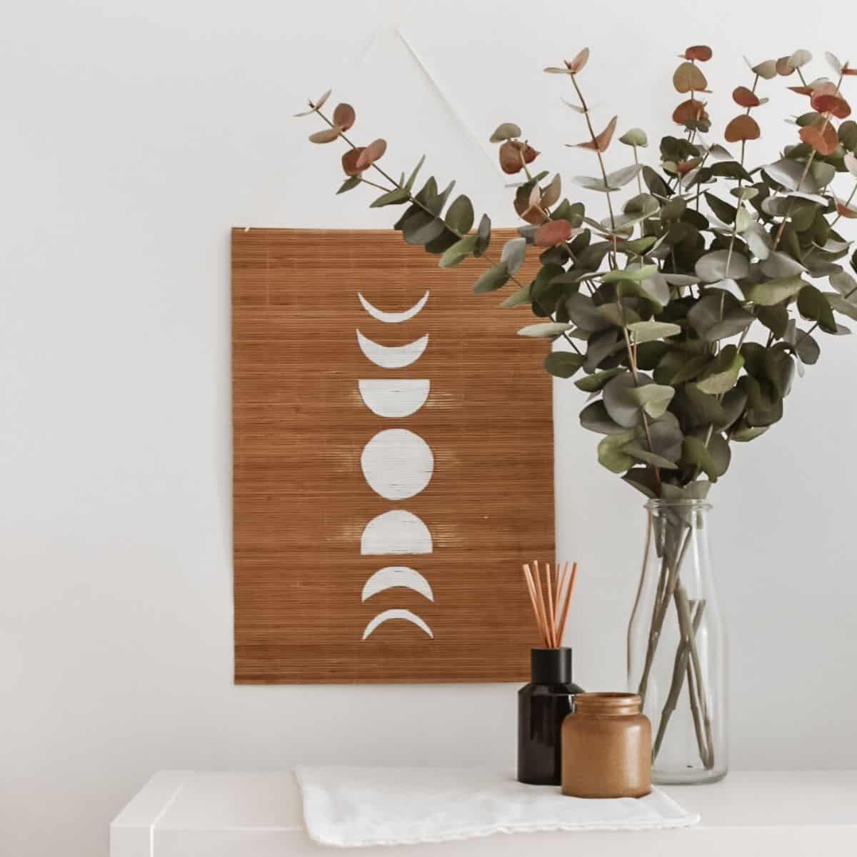 A vase with eucalyptus branches sits on a white table next to a small brown jar and a diffuser. A DIY bamboo wall hanging above beautifully displays the moon phases in white.