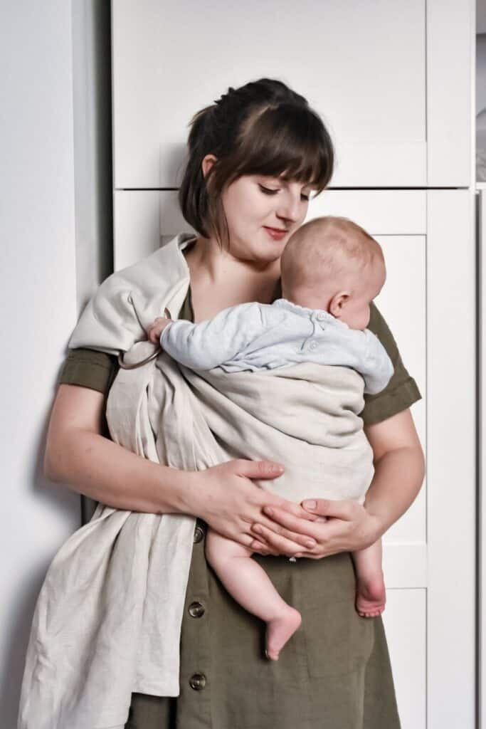 A woman with brown hair cradles a baby in a light-colored DIY linen ring sling against her chest. She is wearing a green dress and smiling softly while standing in a room with a white cabinet in the background.