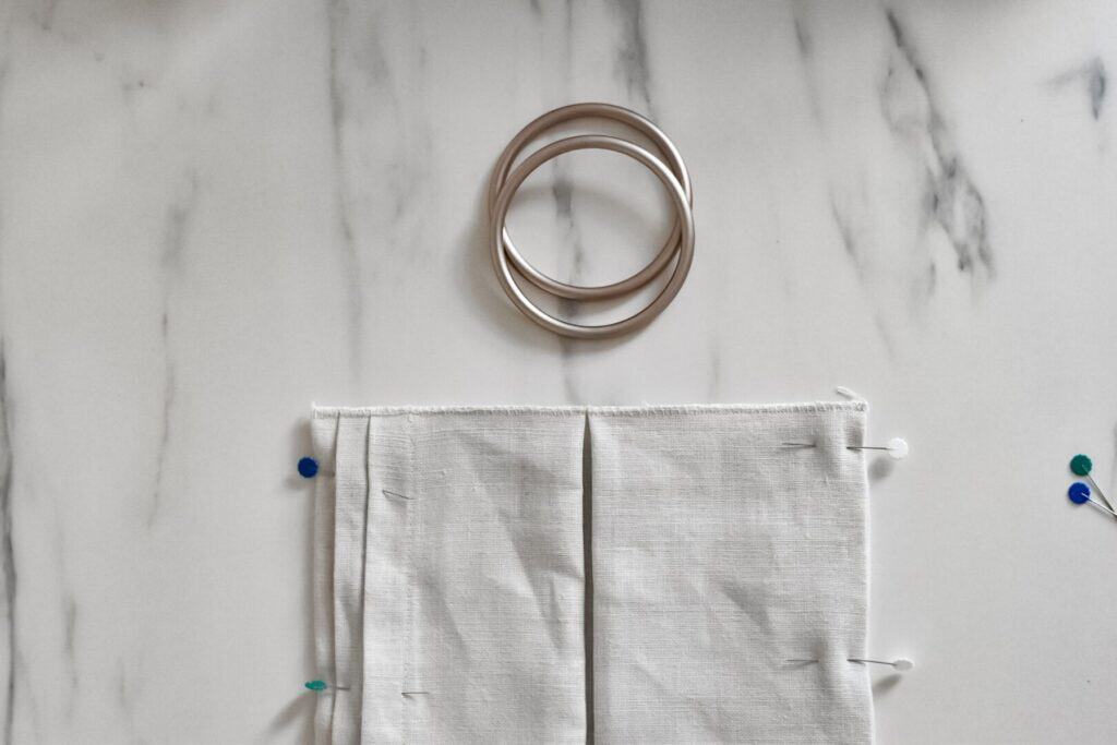 A beige fabric is pinned on a marble surface with two sling rings above it. The raw edge has been finished with an overlocking stitch. A few colorful pins are scattered nearby.