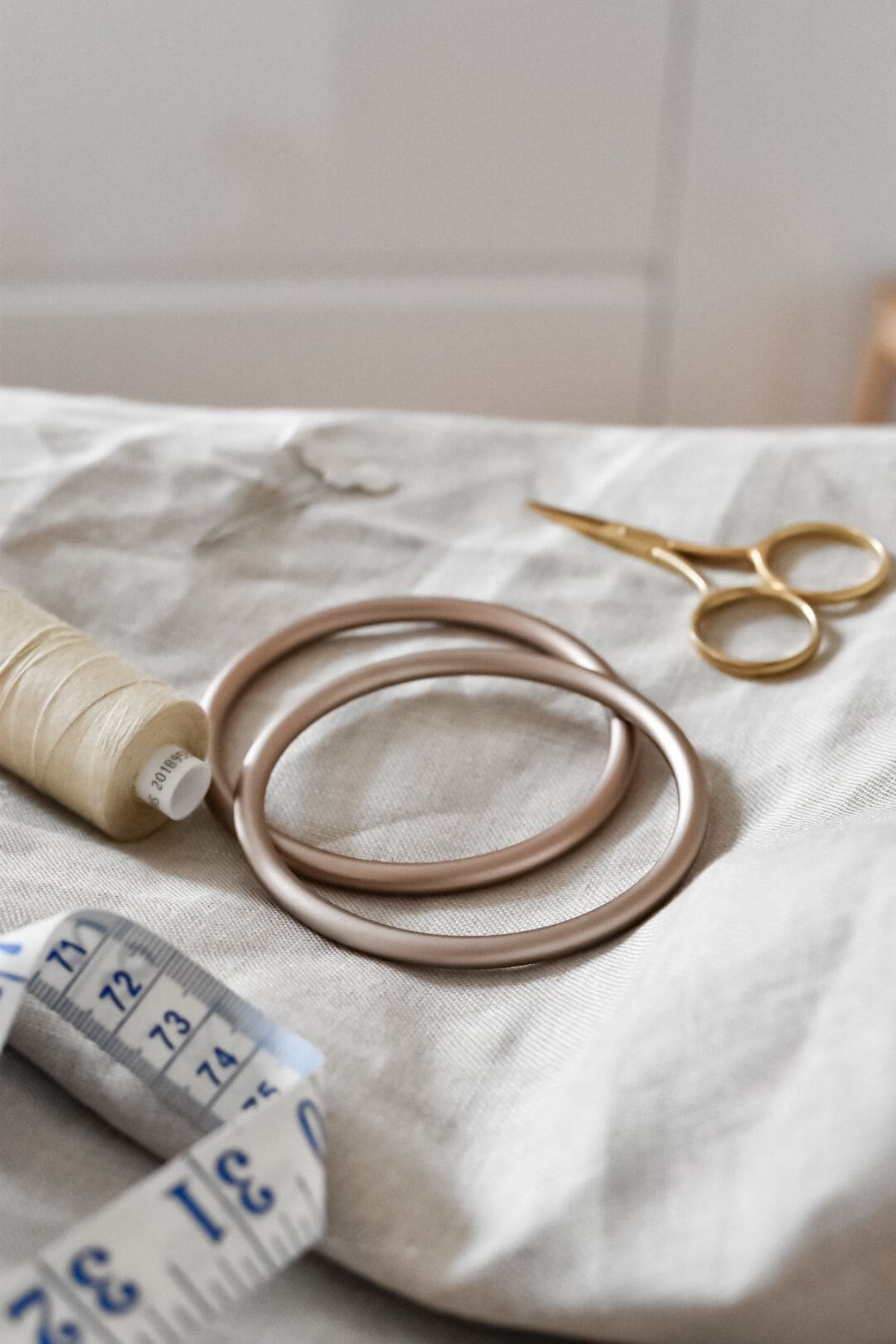 Close-up of sewing tools on a light textile surface, perfect for a DIY project. The image features two sling rings, a spool of cream-colored thread, a pair of gold scissors, and a blue and white measuring tape, ideal for making your own linen ring sling or baby carrier.