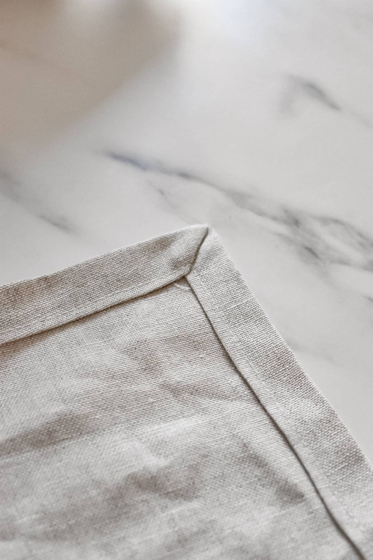 Close-up of a neatly folded corner of a beige linen fabric, ready to make a DIY linen ring sling, placed on a white marble surface. The fabrics texture and the smooth pattern of the marble are visible.