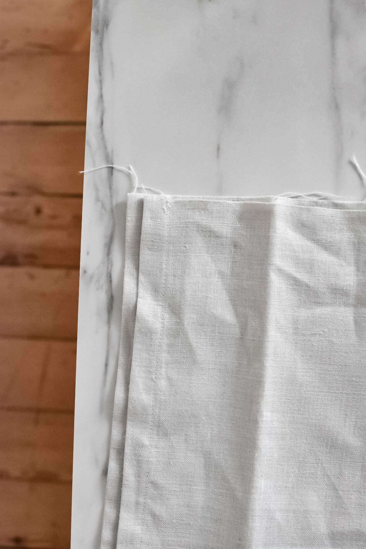 A piece of beige linen with frayed edges is spread on a white marble surface. The two finished edges meet on the left hand side.